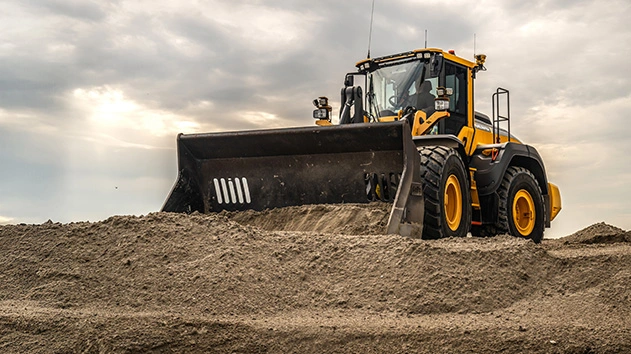 A yellow Volvo wheel loader equipped with Trimble Earthworks grade control moves a large pile of sand under a cloudy sky.