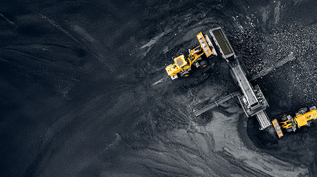An aerial, top-down shot of a large pile of dark, coarse material, likely coal or gravel, with two yellow loaders working.
