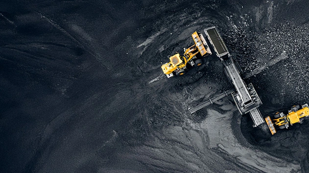 An aerial, top-down shot of a large pile of dark, coarse material, likely coal or gravel, with two yellow loaders working.