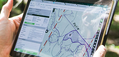 Hands hold a tablet displaying a detailed topographic map with lines and routes in a forest setting, conveying exploration and navigation.