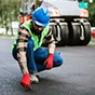 A construction worker in a blue hard hat and red gloves crouches to inspect fresh asphalt near a road roller on a paving site.