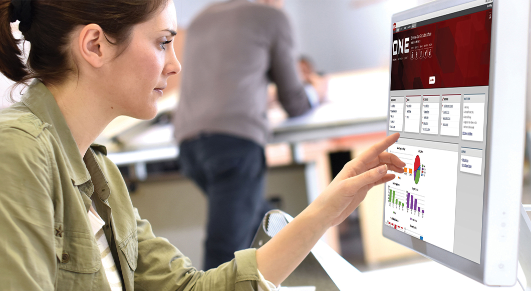 A woman interacts with a Trimble Construction One software dashboard on a touch screen monitor in an office setting.
