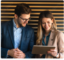 A man and woman in business attire smiling and looking at a tablet together against a horizontal wood-slatted wall.