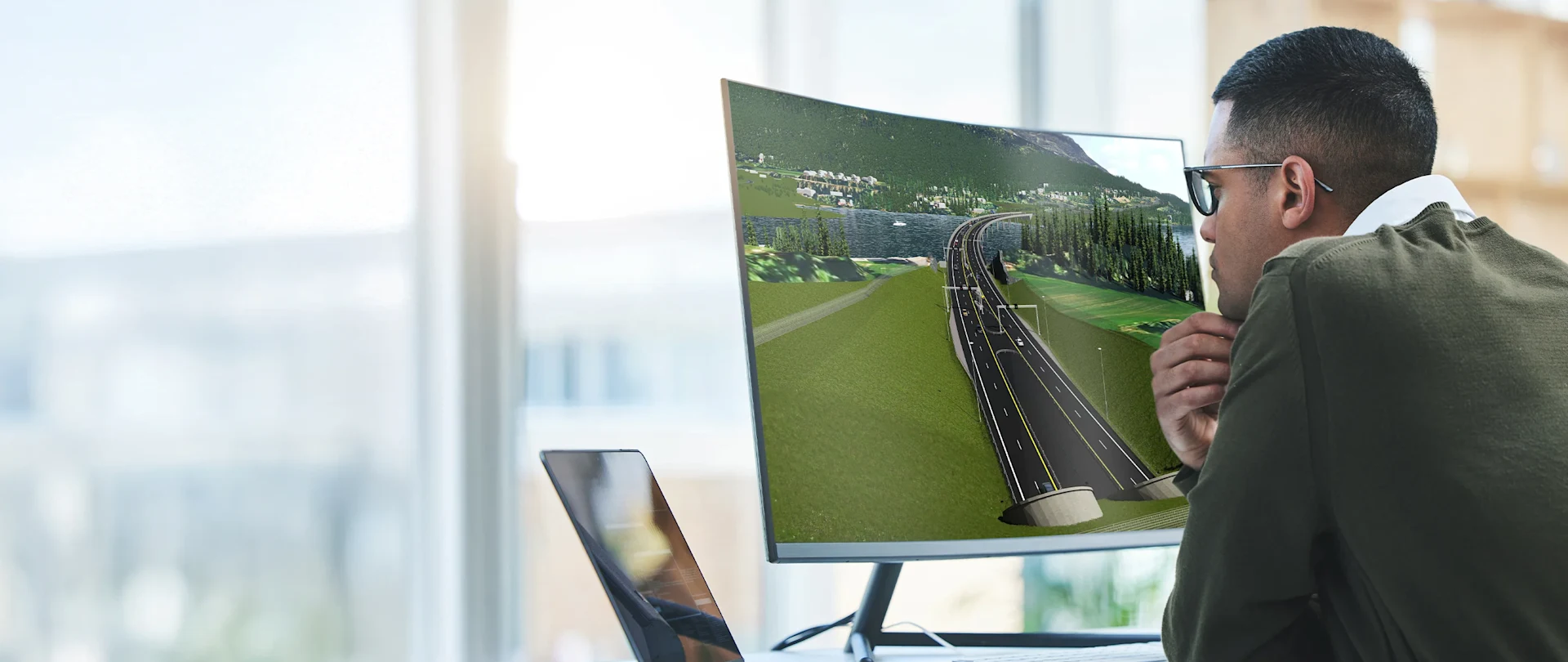 An engineer works at a desk, looking at a 3D road design model on a curved monitor using Novapoint software.