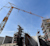 A large yellow tower crane stands over a construction site featuring concrete walls and multi-level scaffolding under a clear blue sky.