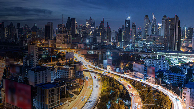An aerial, nighttime long exposure shot captures a bustling highway interchange in a modern city, with the brightly lit roadways winding through a dense urban landscape of tall buildings.