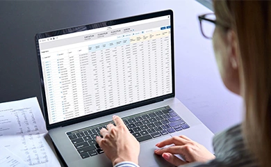 A woman uses a laptop to view a financial spreadsheet and data dashboard within a Trimble software application.