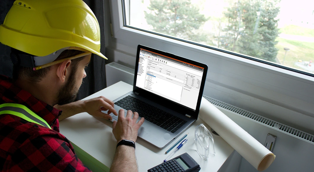 A construction professional in a yellow hard hat uses Trimble construction software on a laptop at a desk with blueprints and a calculator.