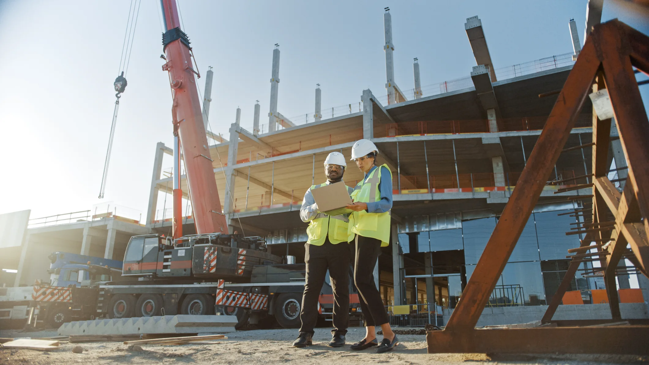 AEC professionals in PPE examine laptop plans at an active concrete construction site. Features: large red mobile crane, partially installed glass facade, structural steel.