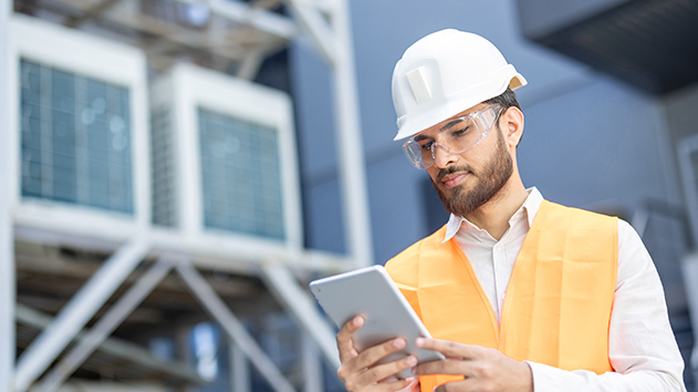 A man in a hard hat and safety glasses looks at a tablet while standing in front of large HVAC units.
