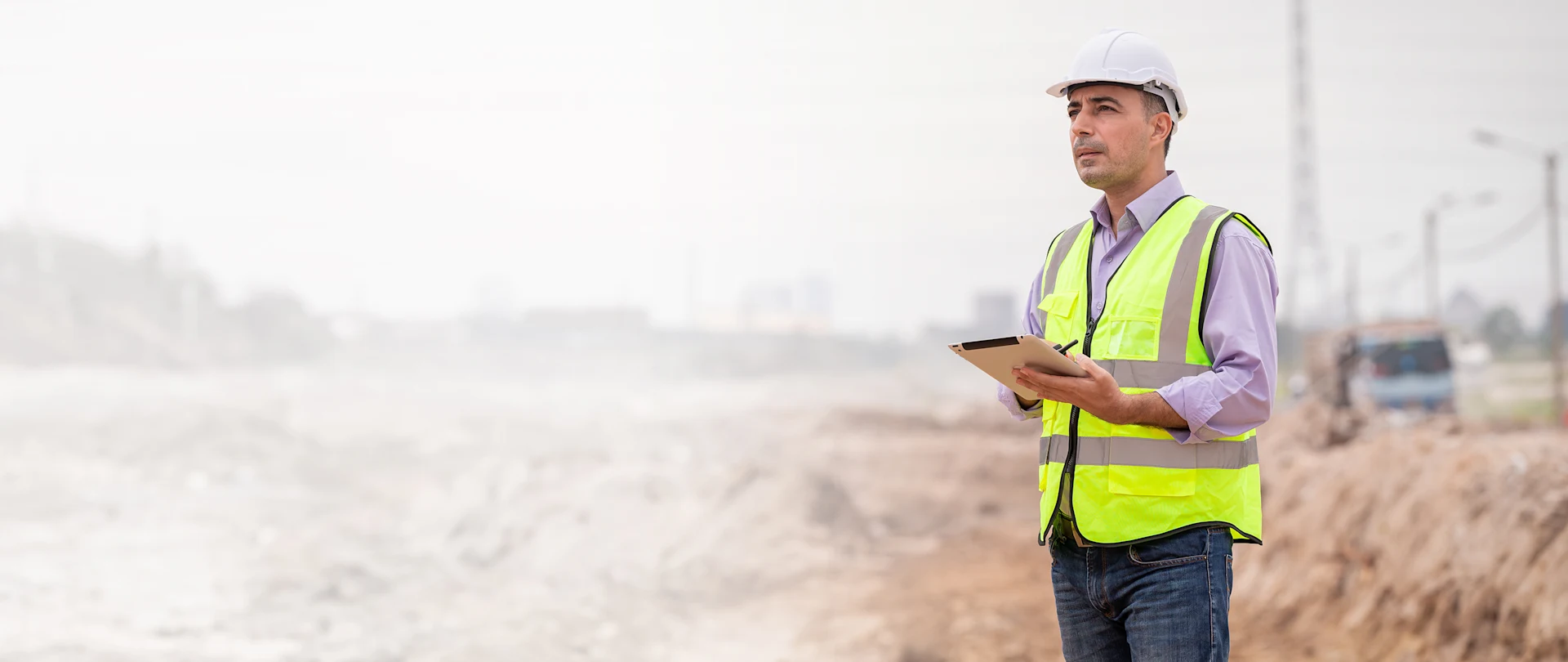 A construction professional in a hard hat and safety vest uses a tablet at a dusty work site.