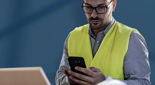 A man in a yellow safety vest and glasses looking at a smartphone, with the corner of a laptop visible in the foreground.