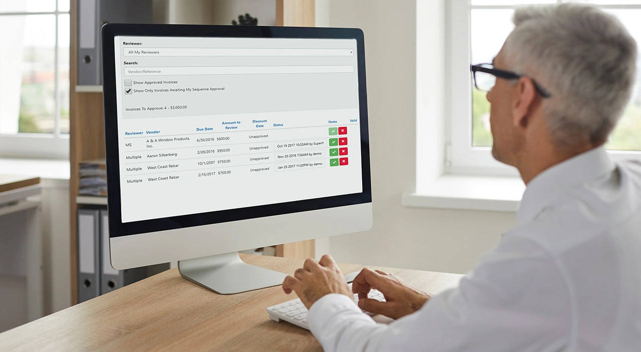 A man uses Trimble Viewpoint Vista software on a computer monitor to review and approve construction invoices.