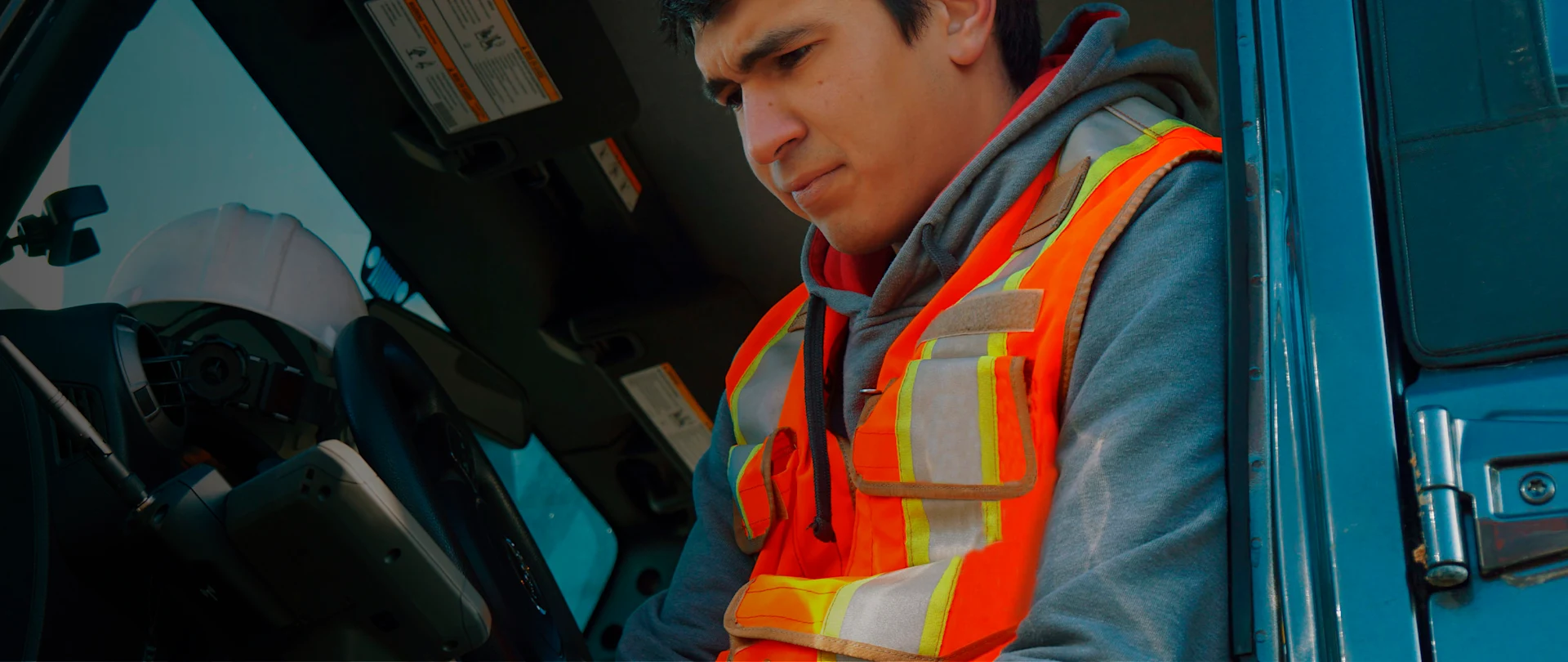 A close-up, eye-level shot shows a man in a gray hooded sweatshirt and a bright orange safety vest with reflective stripes, seated in the cab of a vehicle.