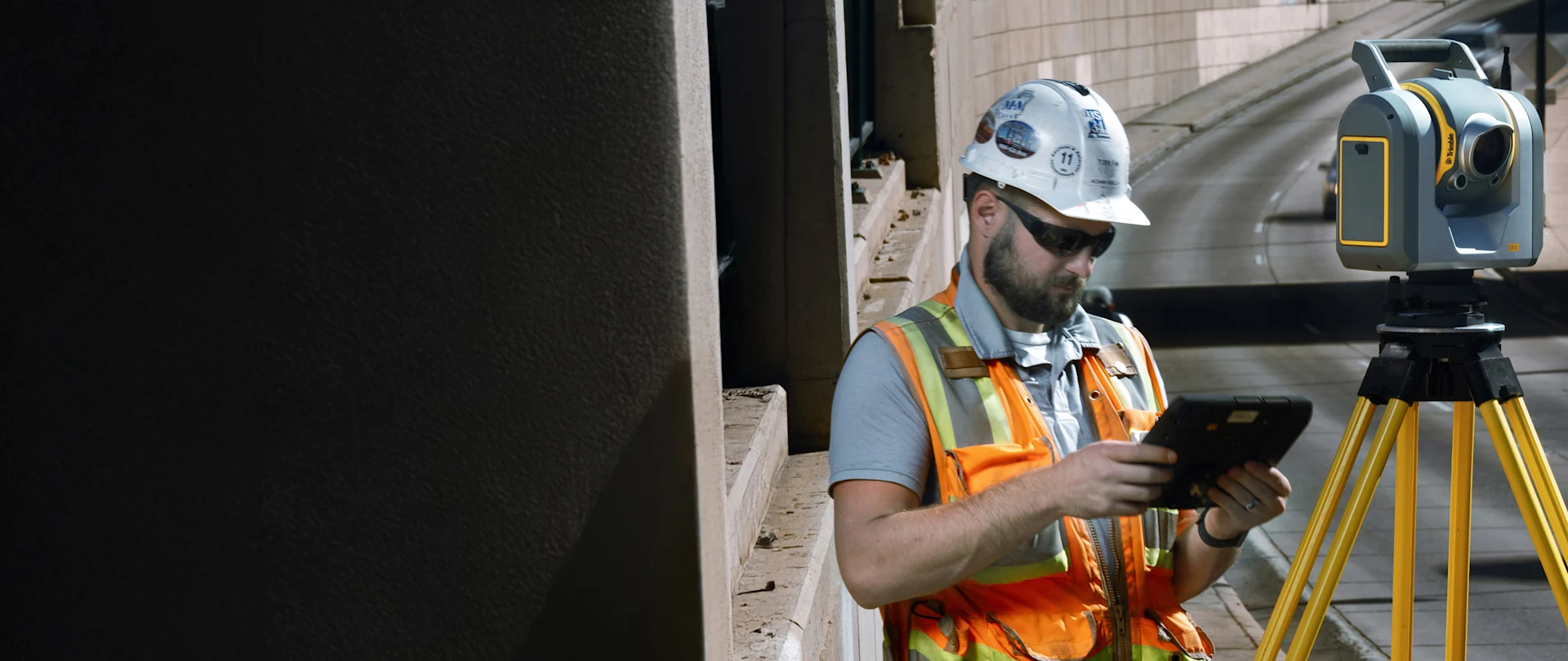 Road engineer in hard hat and safety vest using Trimble SX12 scanner on side of highway with cars driving past.