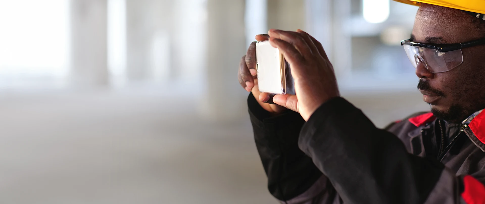 A construction worker in a yellow hard hat and safety glasses uses a smartphone to take a photo inside an open industrial workspace.