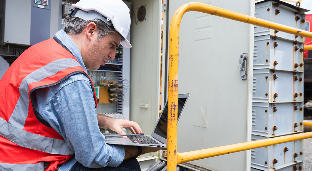 An engineer in a hard hat and safety vest uses a laptop to inspect an industrial electrical control panel on a job site.