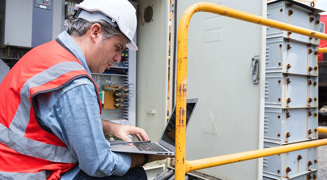An engineer in a hard hat and safety vest uses a laptop to inspect an industrial electrical control panel on a job site.