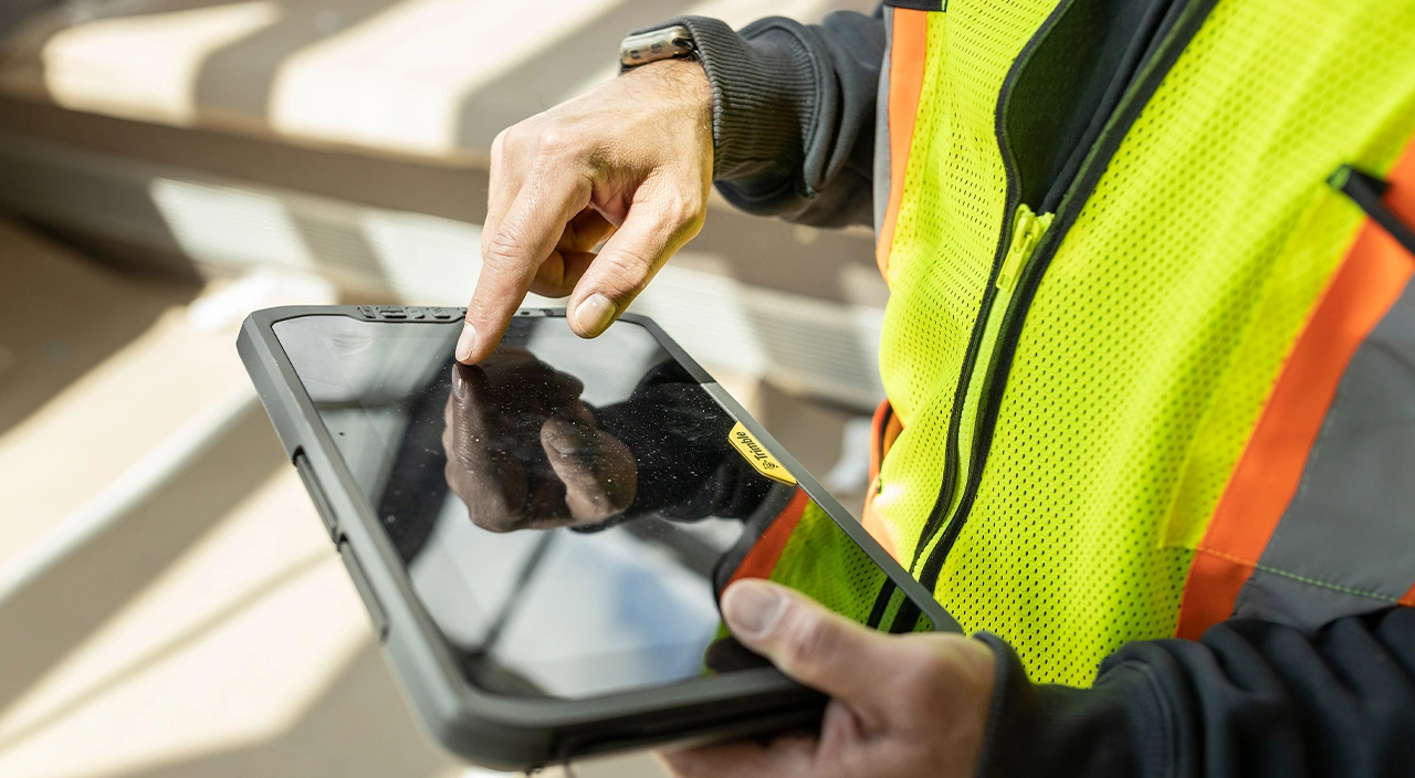 A field worker in a high-visibility safety vest uses a Trimble T100 rugged tablet on a job site.