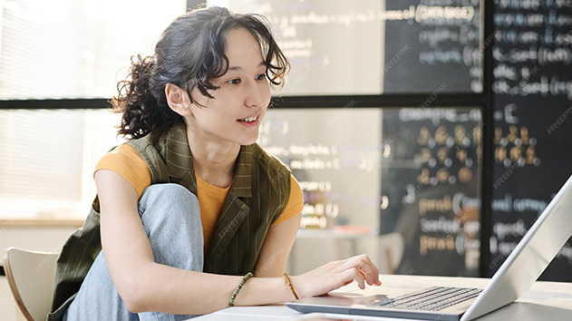 A woman with curly hair smiles while working on a laptop in front of a blackboard covered in Python code.