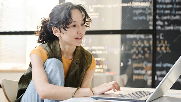 A woman with curly hair smiles while working on a laptop in front of a blackboard covered in Python code.