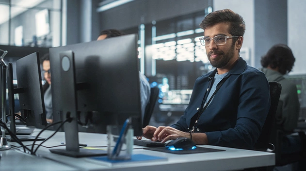 A man with glasses works at a computer in a modern office with multiple screens and people in the background.