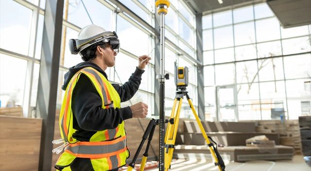 Ein Bauarbeiter auf einer modernen Baustelle, der einen Trimble XR10 mit HoloLens-2-Mixed-Reality-Schutzhelm trägt, nutzt einen R12i-GNSS-Empfänger von Trimble und eine Trimble-Totalstation für Vermessungs- und Absteckarbeiten.