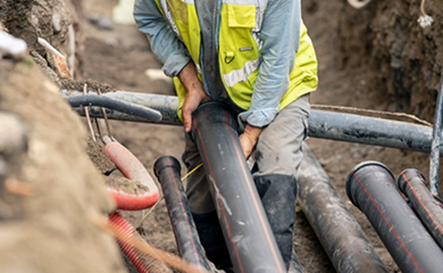 Construction worker in a high-visibility vest installs large black pipes in a trench at a job site.