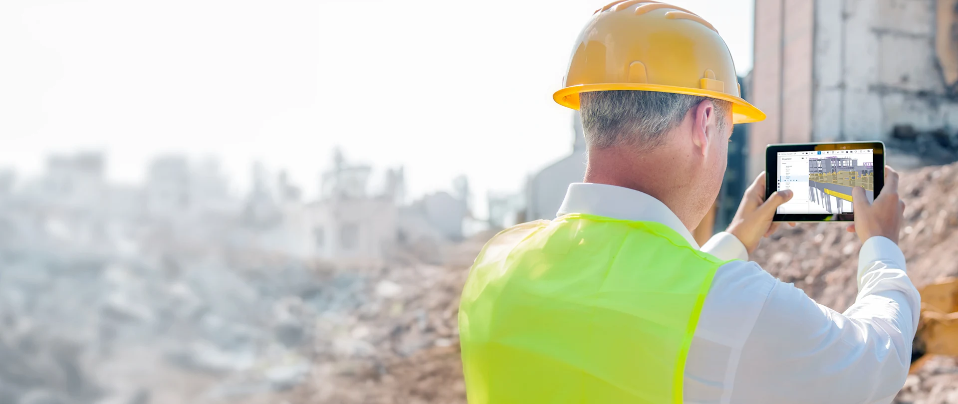 A worker in a yellow hard hat uses a tablet running Trimble Connect software to view a 3D building model at a demolition site.