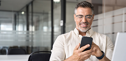 A smiling middle-aged man with glasses and a beard looks at his smartphone in a modern office, wearing a light linen shirt and a black smartwatch.