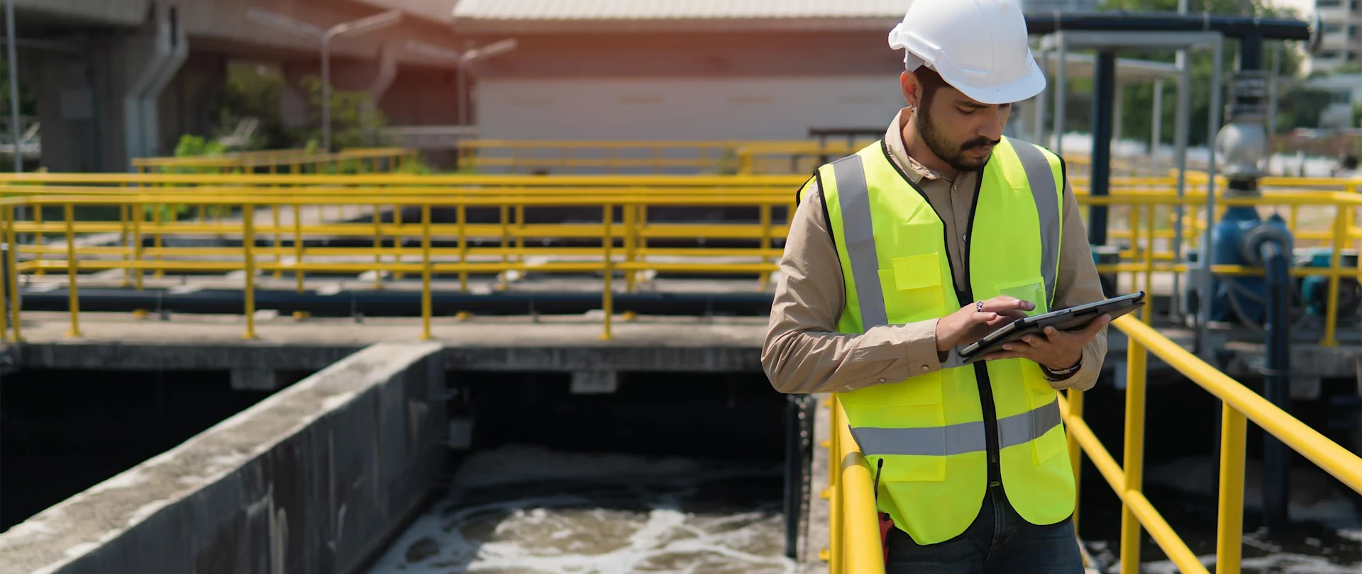 A man in a hard hat and safety vest uses a tablet at a water treatment facility with yellow railings.