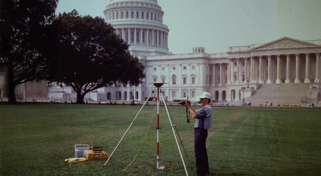 Ein Vermessungsingenieur bedient auf dem Gelände des US-Kapitols ein älteres GPS/GNSS-Vermessungsgerät, das auf einem Stativ mit einer Messstange montiert ist. Daneben stehen ein Datenerfassungsgerät und eine Kühlbox.