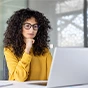 A woman with curly hair and glasses wearing a yellow shirt looks thoughtfully at a laptop in a modern office.