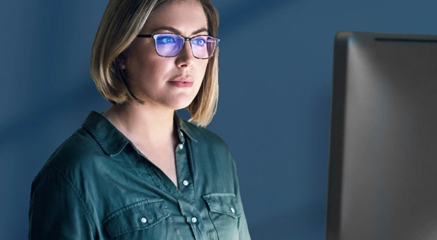 Focused woman with glasses and short blonde hair looking at a computer monitor in a dimly lit room with blue screen light reflections.