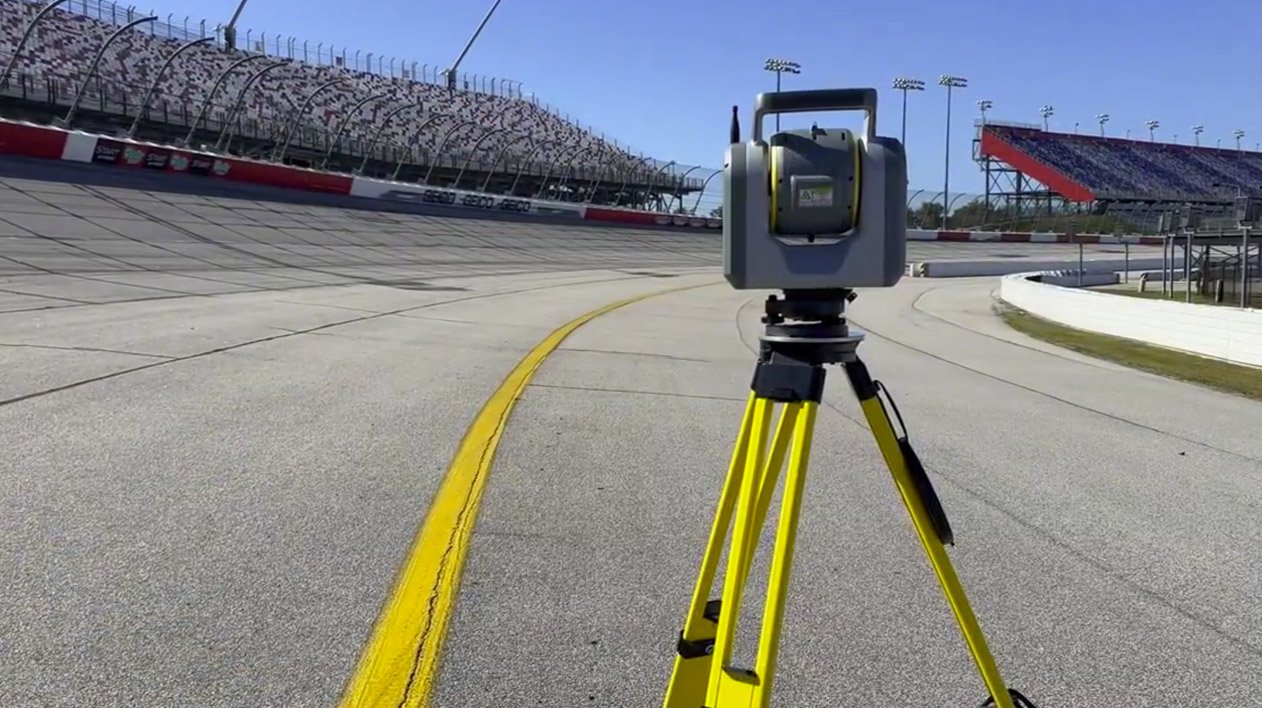 A Trimble 3D laser scanner on a yellow tripod is set up on at the Darlington race track with empty grandstands visible in the background.