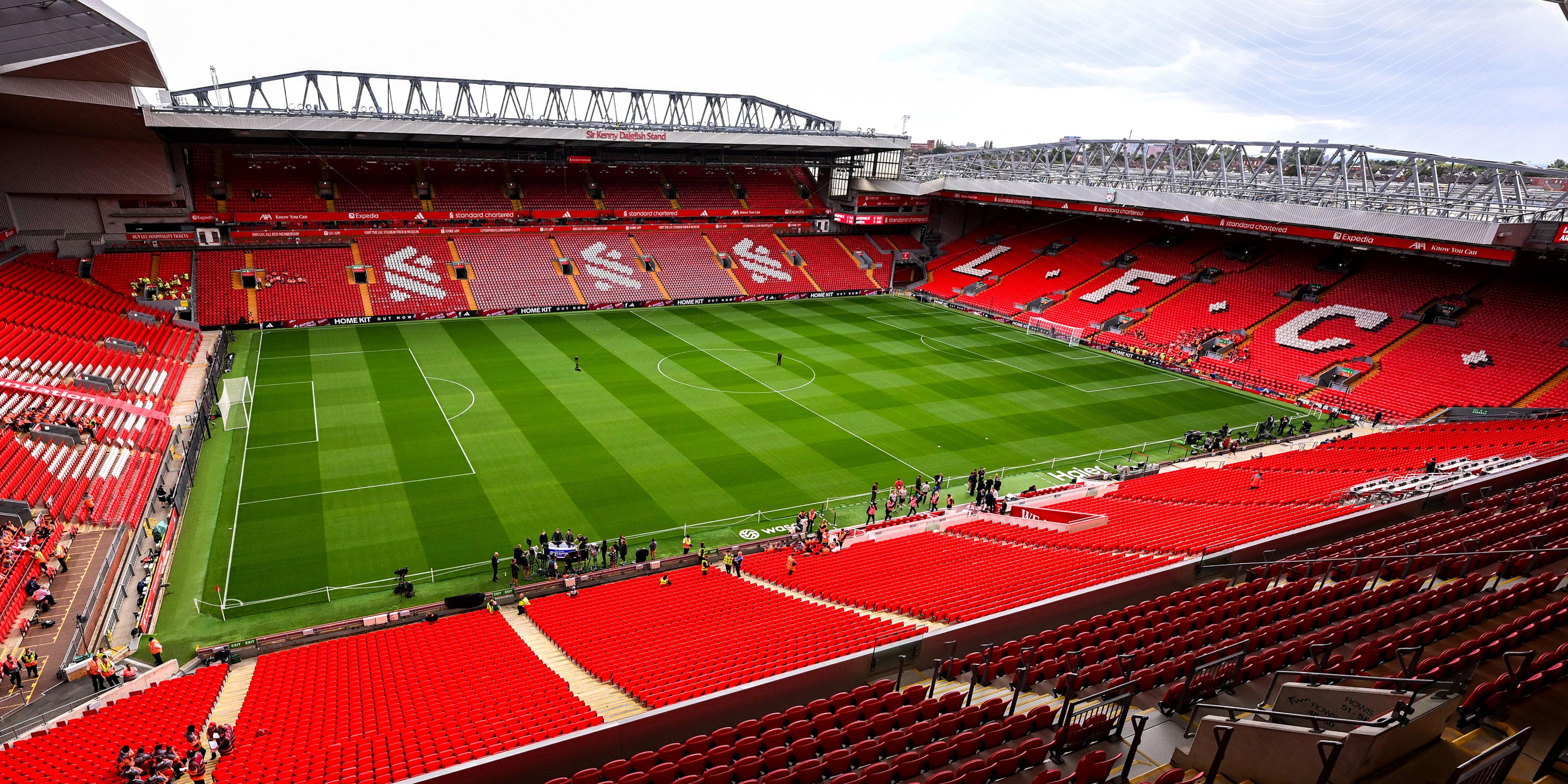 Anfield stadium empty during the day, showing vivid red stands with "LFC" spelled in white seats, a green pitch, and modern roof structures.