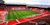 Anfield stadium empty during the day, showing vivid red stands with "LFC" spelled in white seats, a green pitch, and modern roof structures.
