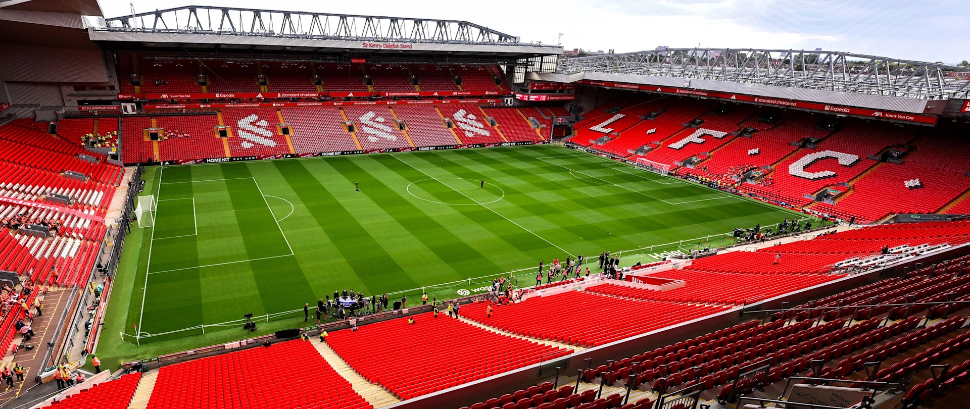 Anfield stadium empty during the day, showing vivid red stands with "LFC" spelled in white seats, a green pitch, and modern roof structures.