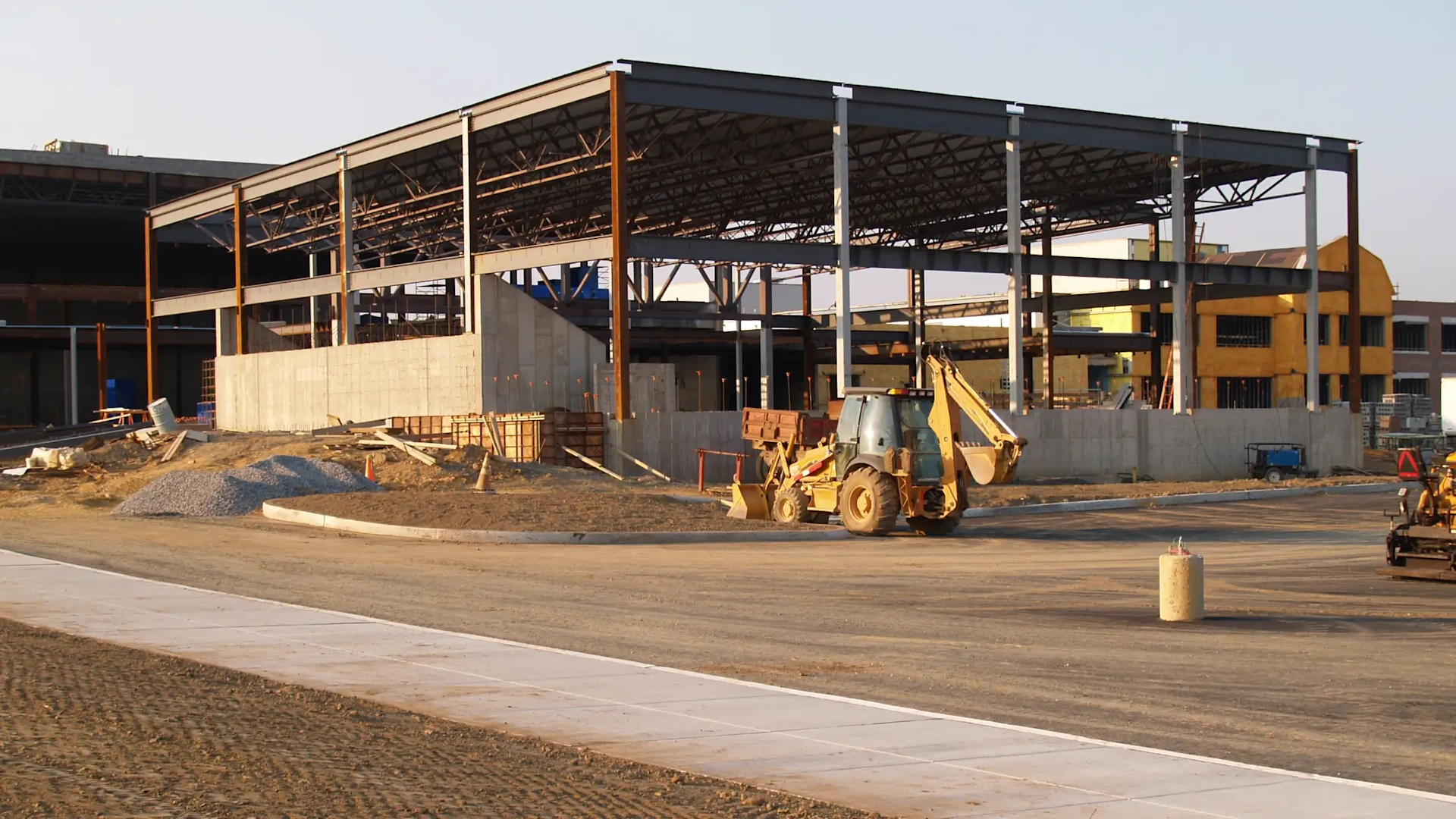 Active AECO construction site: steel-framed building, concrete foundations, yellow backhoe loader, aggregate piles, formwork, and new concrete pavement.