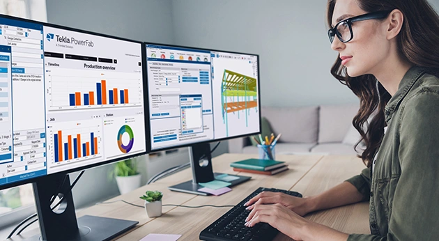 A woman uses Trimble Tekla PowerFab software on dual monitors to view steel fabrication data and a 3D structural model.