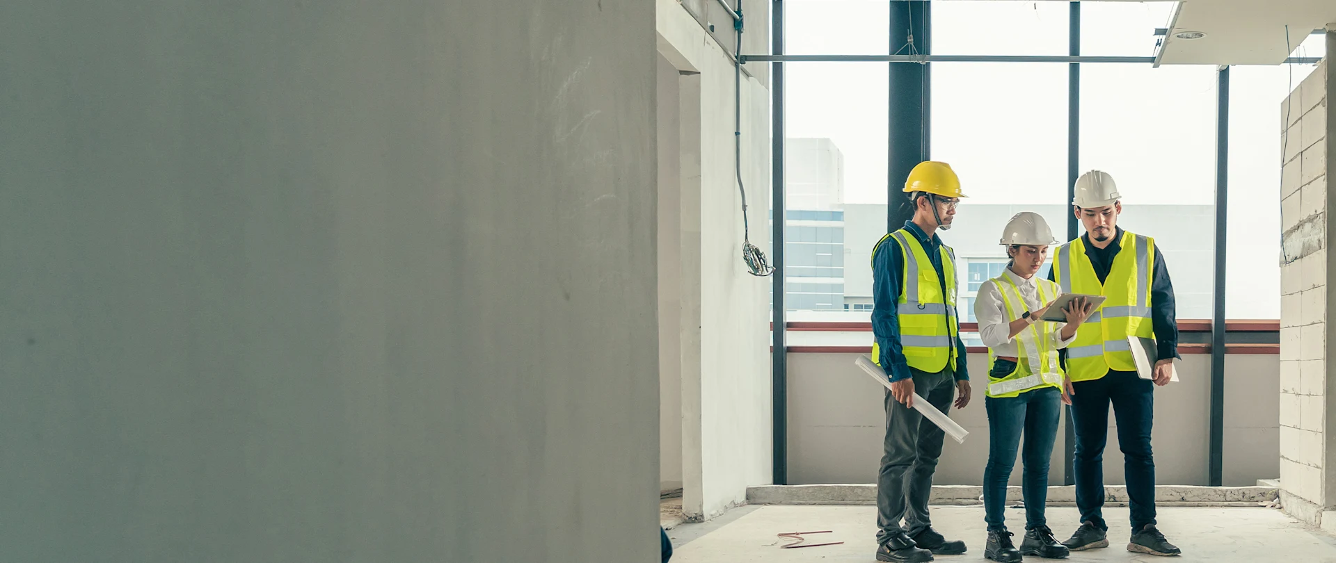 Three construction professionals in hard hats and safety vests review plans on a tablet and laptop in an unfinished building.
