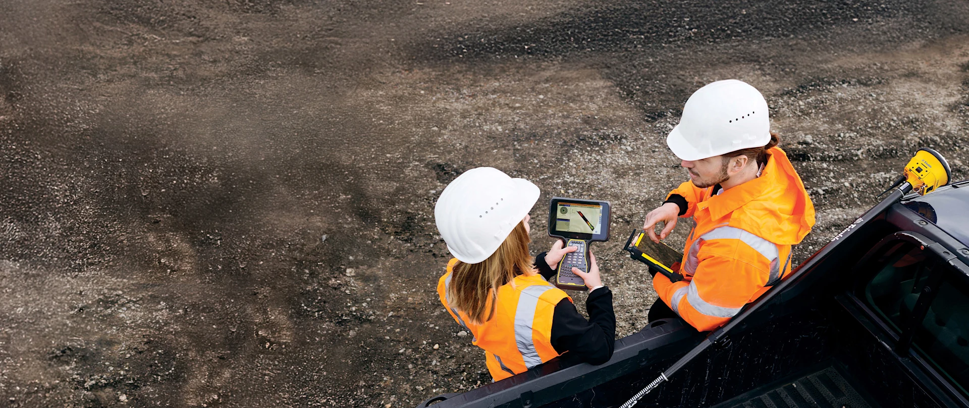 Two construction workers in hard hats and vests look at a handheld device while leaning on a truck.