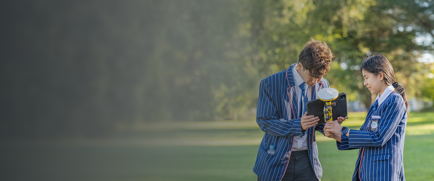 Two individuals in striped blazers are standing outdoors on a grassy field, examining a device mounted on a tripod.