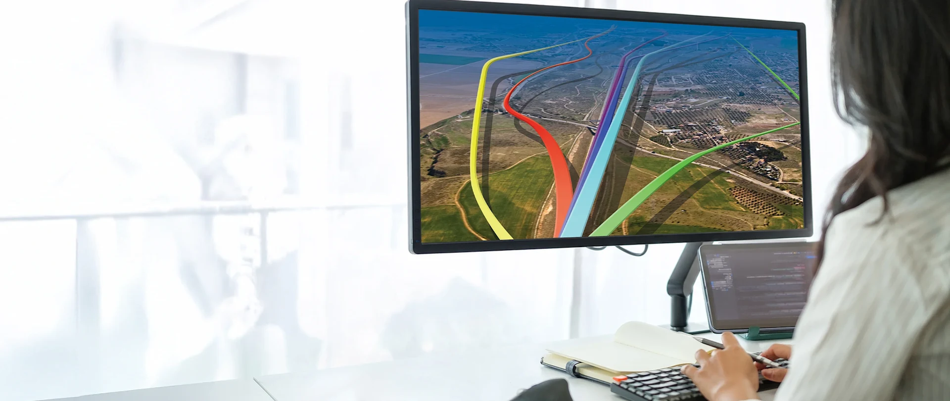 A person works at a computer using Quantm software, displaying colorful 3D route alignment paths over a 3D landscape terrain.