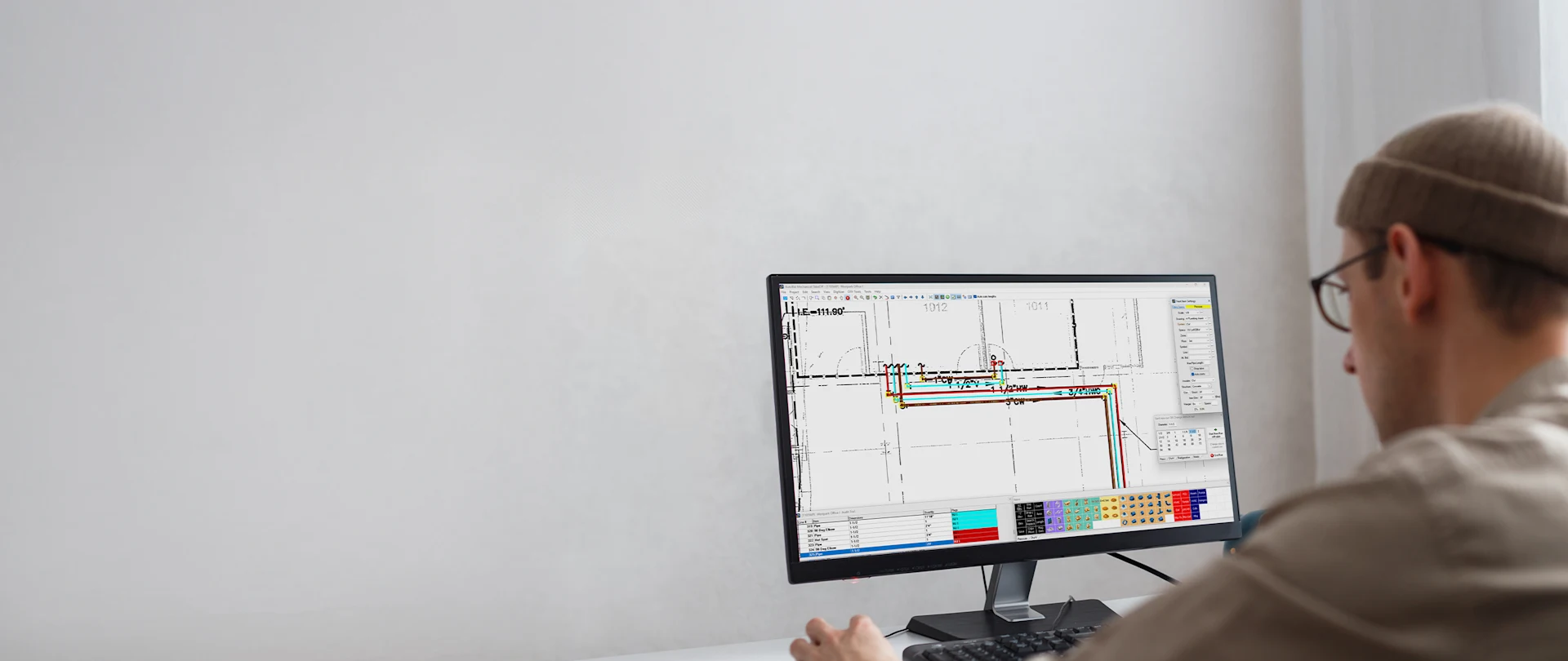 A person wearing a beanie sits at a desk, looking at a computer screen displaying Trimble AutoBid Mechanical software.