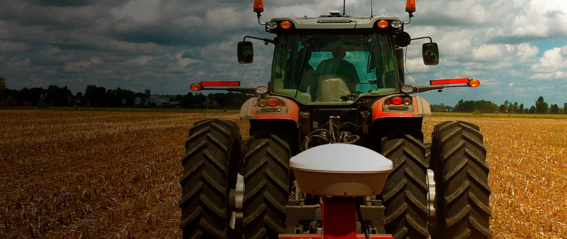 A rear-view, low-angle shot captures a modern tractor in a field of harvested crops under a dramatic, cloudy sky.