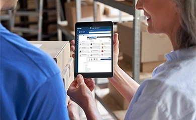 Two workers in a warehouse use a tablet to access Trimble's TRA-SER software for product information and pricing.