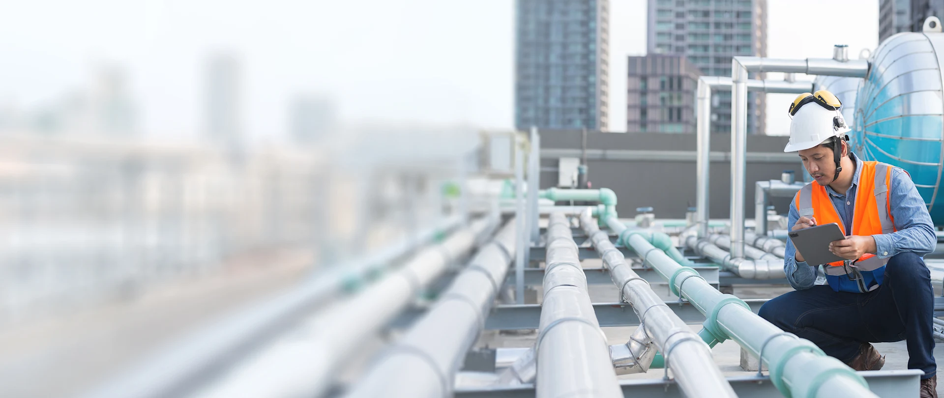 An engineer in a safety vest uses a tablet to inspect industrial pipes on a city rooftop, representing Trimble MEP connected workflows.