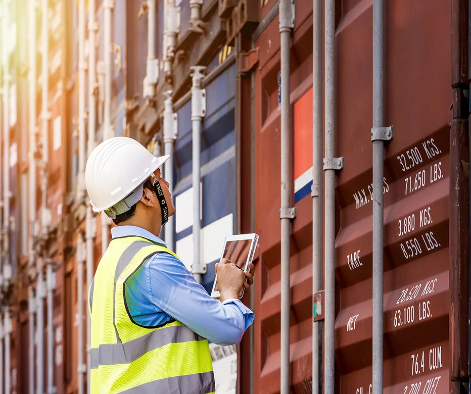 Man with reflective vest and hard hat on tablet inspecting a shipping container