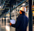 Een MEP-professional met een witte veiligheidshelm kijkt omhoog naar complexe mechanische, elektrische en loodgieterswerk (MEP) leidingen in een industriële faciliteit, terwijl hij een tablet met een datadashboard vasthoudt.